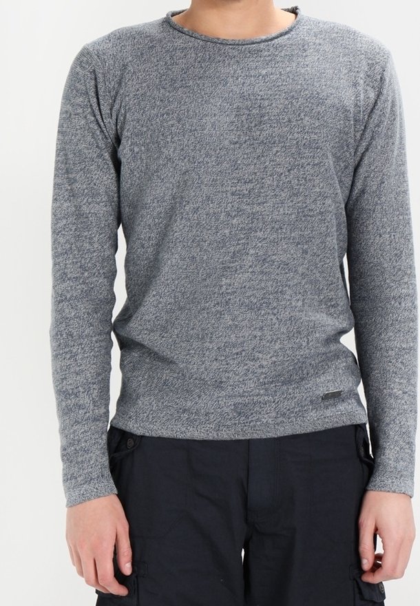 Homme portant un pull gris ajusté à manches longues en maille et un pantalon cargo noir, debout devant un fond clair uni.
