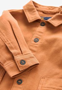 Rostfärgad manchester skjorta med krage, långa ärmar och mörka knappar. Framträdande ribbad textur med en bröstfickdetalj.