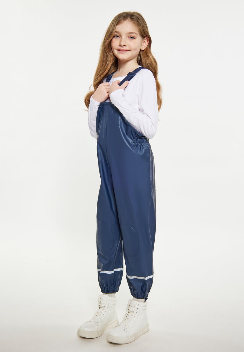 Jauna mergaitė, vilkinti tamsiai mėlynus vandeniui atsparius kombinezonus ir baltą ilgomis rankovėmis marškinį, stovi ir laiko kombinezono dirželius abiem rankomis.