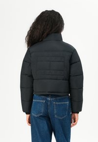 Black cropped puffer jacket with a high collar, quilted texture, and embroidered "TOMMY JEANS" logo on the back. Paired with blue jeans.