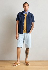 Chemise à manches courtes bleu marine boutonnée sur un t-shirt moutarde, associée à un short en denim clair et des sandales marron. Comprend un foulard bleu autour du cou.