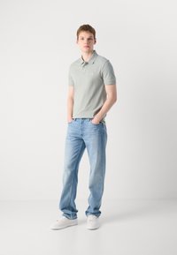Lysegrønn polo skjorte med knappeskrage, sammen med lyseblå rette jeans og hvite joggesko. Modellen står med hendene i lommer.