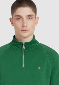 Green zip-up sweatshirt made of soft fabric, featuring a high collar and orange logo on the chest. Smooth texture and minimalistic design.