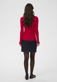 Frau mit langen welligen braunen Haaren, die ein rotes Langarmoberteil, einen dunklen Jeansrock, schwarze Strumpfhosen und schwarze Slipper trägt und mit dem Rücken zugewendet steht.