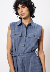 Ärmelloses Jeanskleid mit vertikalen weißen Streifen, zwei vorderen Taschen, Knopfdetails und einem gebundenen Taillengürtel.