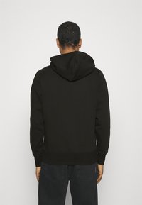 Man vänd bort från kameran, klädd i svart luvtröja och mörka jeans, står mot en enkel ljus bakgrund.