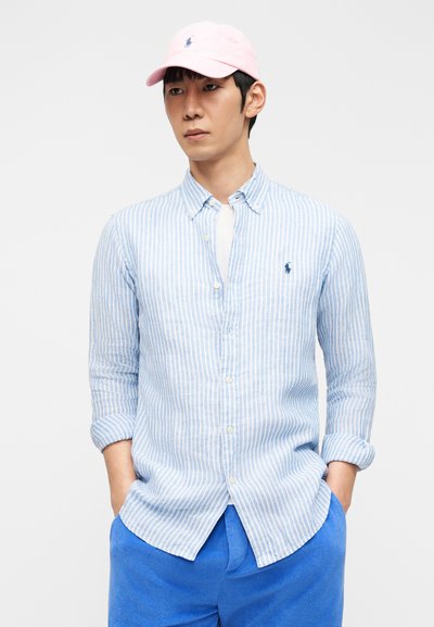 Άντρας που φοράει ροζ καπέλο, γαλάζιο και λευκό ριγέ πουκάμισο με κουμπιά και λογότυπο, λευκό εσώρουχο και φωτεινό μπλε παντελόνι, με τα χέρια στις τσέπες.