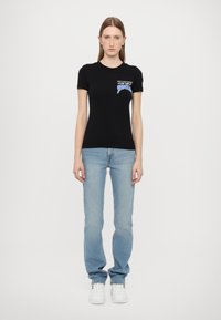 T-shirt nera a maniche corte con una piccola grafica colorata sul lato sinistro del petto, abbinata a jeans blu chiaro e sneakers bianche.