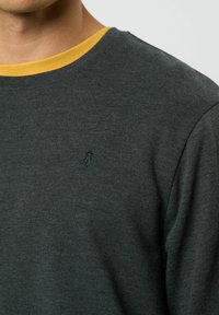 Sötét szürke pulóver kerek nyakkal és sárga belső gallérral. Kises hímzett kék logóval a mellrészen. Puha tapintású.