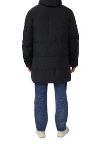 Manteau rembourré noir, avec capuche, design matelassé à lignes horizontales, et coupe longue, associé à un jean bleu et des chaussures légères.