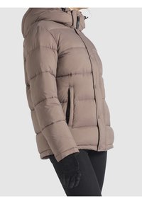 Chaqueta marrón acolchada con cierre de cremallera, capucha y bolsillos laterales. Presenta un diseño acolchado y puños elásticos. Se llevan guantes negros.