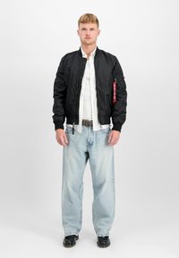 Chaqueta bomber negra con puños y cintura acanalados, combinada con una camisa blanca y jeans holgados de color azul claro. Detalle de bolsillo notable en la manga.