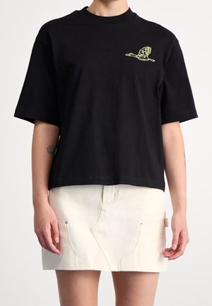 Persona con una camiseta negra de gran tamaño con un pequeño logo amarillo de pájaro y una falda corta color marfil con bolsillos delanteros.