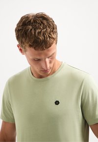 Lichtgroen T-shirt met ronde hals, gemaakt van zachte stof, met een klein zwart zeshoekig logo op de linkerborst. Soepele textuur, casual ontwerp.