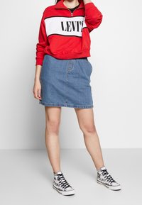 Rode en witte ritstrui met het "LEVI'S" logo, gecombineerd met een spijkerrok en zwart-witte hoge sneakers met grafisch ontwerp.