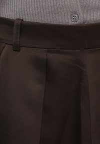 Pantaloni marroni plissettati con una texture liscia e lucida e una cintura con passanti. Abbinati a un cardigan grigio a costine con dettaglio di bottoni.