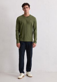 Długorękawowa koszulka w kolorze oliwkowej zieleni wykonana z miękkiego materiału, z małym logo na piersi. Połączona z granatowymi spodniami i białymi tenisówkami.