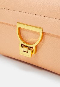 Szczegół torebki ze skóry ziarnistej w kolorze brzoskwiniowym z błyszczącym złotym zapięciem z grawerem "COCCINELLE" na białym tle.