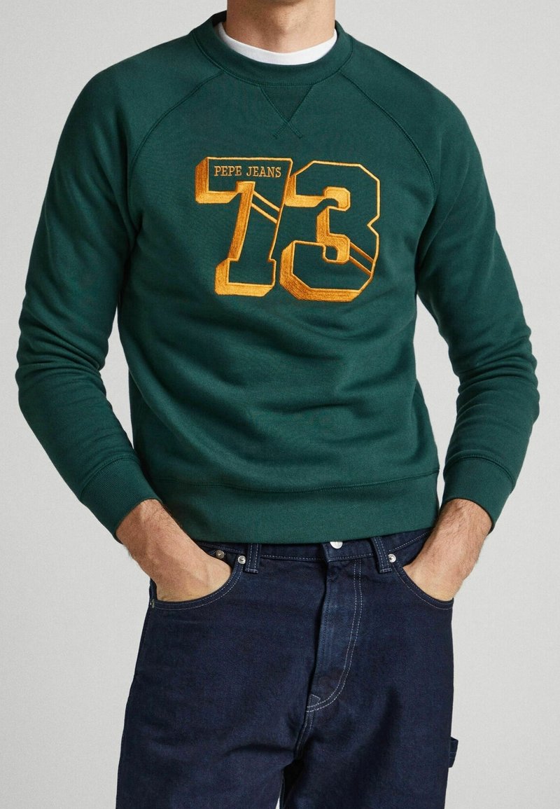 Homme portant un sweat-shirt vert foncé avec le texte jaune "PEPE JEANS 73" et un jean bleu foncé, les mains dans les poches, sur un fond uni.