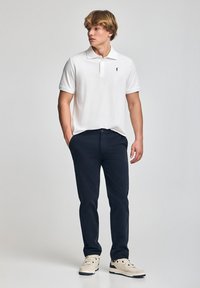Biała koszulka polo z czarnym logo, zestawiona z granatowymi spodniami i jasnymi sneakersami. Tkanina wydaje się gładka i ma klasyczny krój.