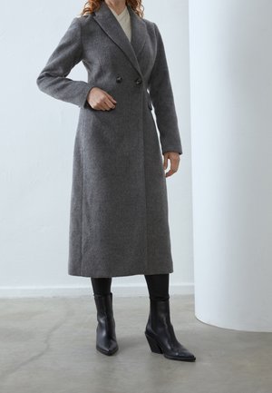 Femme portant un long manteau gris à double boutonnage, des leggings noirs et des bottines noires à talons, debout à l'intérieur sur un sol gris.