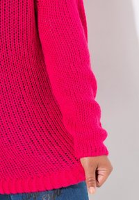 Różowy sweter z dzianiny o luźnym splotcie i prążkowanych mankietach, z charakterystycznym wzorem i zaokrąglonym dołem. Widoczna dłoń trzymająca materiał.