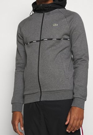 Uomo che indossa una felpa con zip grigia con cappuccio nero e banda orizzontale nera con scritto "Lacoste", abbinata a pantaloni neri con strisce rosse e bianche.