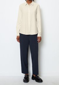 Camisa beige de botones de manga larga con cuello, combinada con pantalones sueltos azul marino y zapatos de plataforma negros. Tela suave, diseño minimalista.