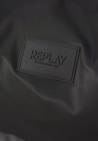 Superficie in nylon nero con una toppa rettangolare in pelle nera in rilievo, con la scritta "REPLAY" e "ESTABLISHED 1981" in lettere grasse.