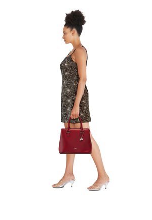 Frau in einem dunklen ärmellosen Kleid mit Blumenmuster geht, hält eine rote Handtasche, trägt transparente Sandalen mit Absatz, schaut nach links.