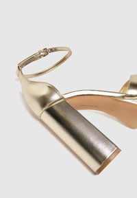 Złote metaliczne sandały na wysokim obcasie z grubym obcasem, paskiem na kostkę, gładką fakturą i otwartym noskiem. Widoczny element ozdobny na pasku.