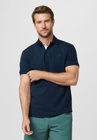 Uomo con capelli corti castani, indossa una polo blu scuro e pantaloni verdi, che aggiusta la manica sinistra davanti a uno sfondo chiaro e uniforme.