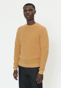 Pull en tricot jaune moutarde avec un col rond, des manches longues et des poignets côtelés, associé à un jean foncé. Texture lisse, coupe ajustée.