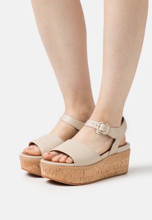 Piedi che indossano sandali open-toe beige con cinturini alla caviglia e tacchi in sughero contro uno sfondo bianco.