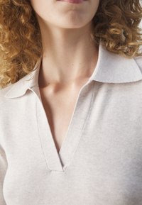 Personne aux cheveux bouclés portant un haut en maille beige clair avec un col ouvert large et un décolleté en V, montrant une partie du cou et du menton.