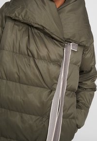 Olivgrön vadderad jacka med texturerad yta. Har en bred krage och ett dekorativt band med fiskbensmönster framtill.