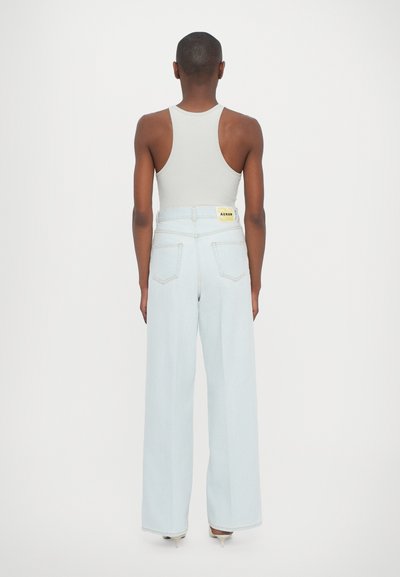 Jeans de ganga de lavagem clara com corte largo, cintura alta e bolsos traseiros. Apresenta uma etiqueta amarela na cintura e uma textura suave.