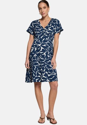 Femme portant une robe bleu marine mi-longue avec des motifs abstraits de feuilles blanches, manches courtes et col en V, debout et faisant face à l'avant.