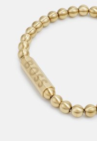Pulsera de cuentas de oro que presenta cuentas redondeadas y un cierre cilíndrico grabado con "BOSS". Textura suave con un acabado mate.