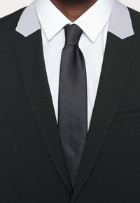 Zwarte colbert met grijze kraagaccenten, wit shirt en zwarte satijnen stropdas. Materiaal lijkt textuur te hebben; op maat gemaakte pasvorm met revers ontwerp.