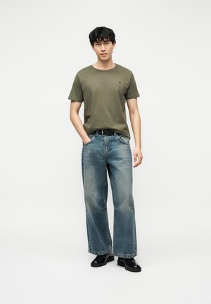 Ung mann står med hendene i lommene, iført olivengrønn t-skjorte, vide, falmede jeans, svart belte og svarte sko mot hvit bakgrunn.