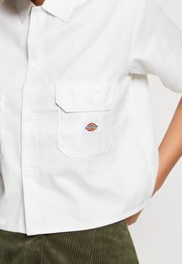 Chemise blanche à manches courtes en tissu lisse, avec deux poches avant et un écusson coloré sur la poche gauche.