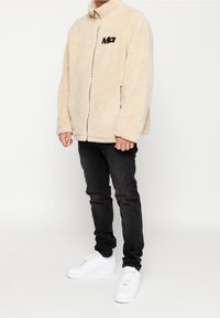 Beżowa polarowa kurtka zapinana na zamek z wysokim kołnierzem, czarnym logo na piersi, zestawiona z ciemnymi dżinsami i białymi sneakersami. Miękka faktura i luźny krój.