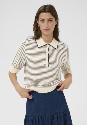 Nő, aki krémszínű, fekete csíkos rövid ujjú galléros inget és sötétkék, réteges szoknyát visel, egyszerű háttér előtt áll.