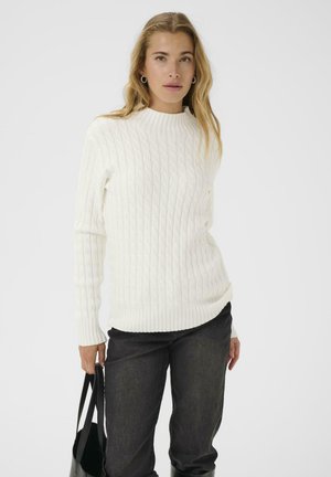 Pull en tricot côtelé blanc avec un col montant et des manches longues, associé à un jean noir et un sac noir. Texture lisse et design ajusté.