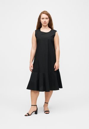 Vrouw met lang rood haar, gekleed in een mouwloze zwarte midi-jurk en zwarte open-toe hakken, staand tegen een effen witte achtergrond.