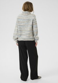 Kobieta stoi tyłem, ubrana w gruby sweter z golfem w odcieniach szaro-niebieskich, czarne szerokie spodnie oraz baleriny w zebrze.