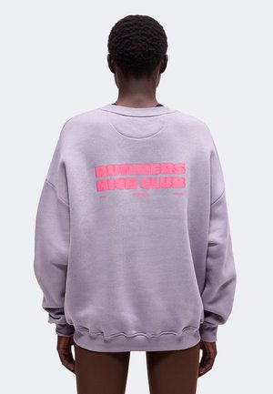 Personne aux cheveux courts et bouclés portant un sweat-shirt violet clair avec un texte rose "RUNNERS HIGH CLUB" dans le dos, debout devant un fond uni.