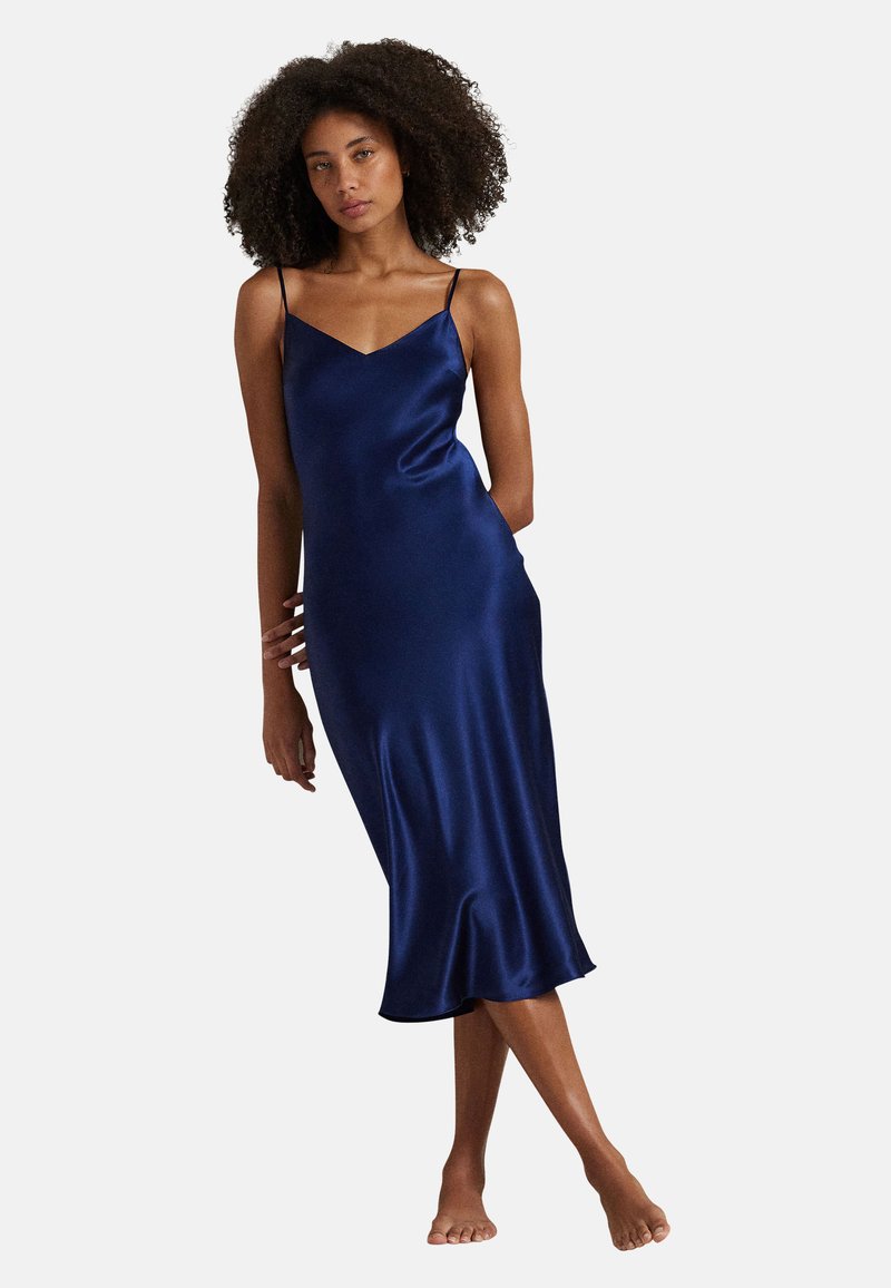 Vrouw met krullend haar die een mouwloze marineblauwe satijnen midi-jurk draagt, blootsvoets staand tegen een effen lichte achtergrond.