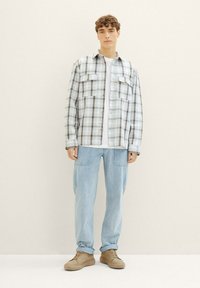 Junger Mann, der ein kariertes Hemd in Weiß und Grau über einem weißen T-Shirt, hellblaue Jeans und beige Sneaker trägt, vor einem schlichten hellen Hintergrund steht.